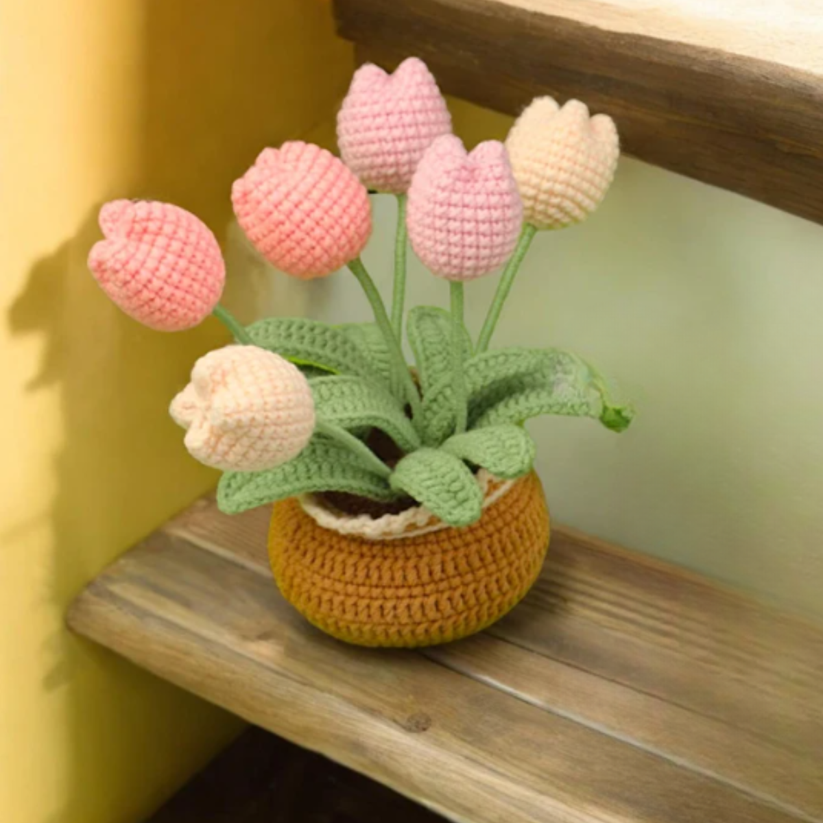 A brown crochet pot made with the Crochet Tulip Kit holds six pink and cream tulips with green stems and leaves, displayed on a wooden step beside a yellow wall.