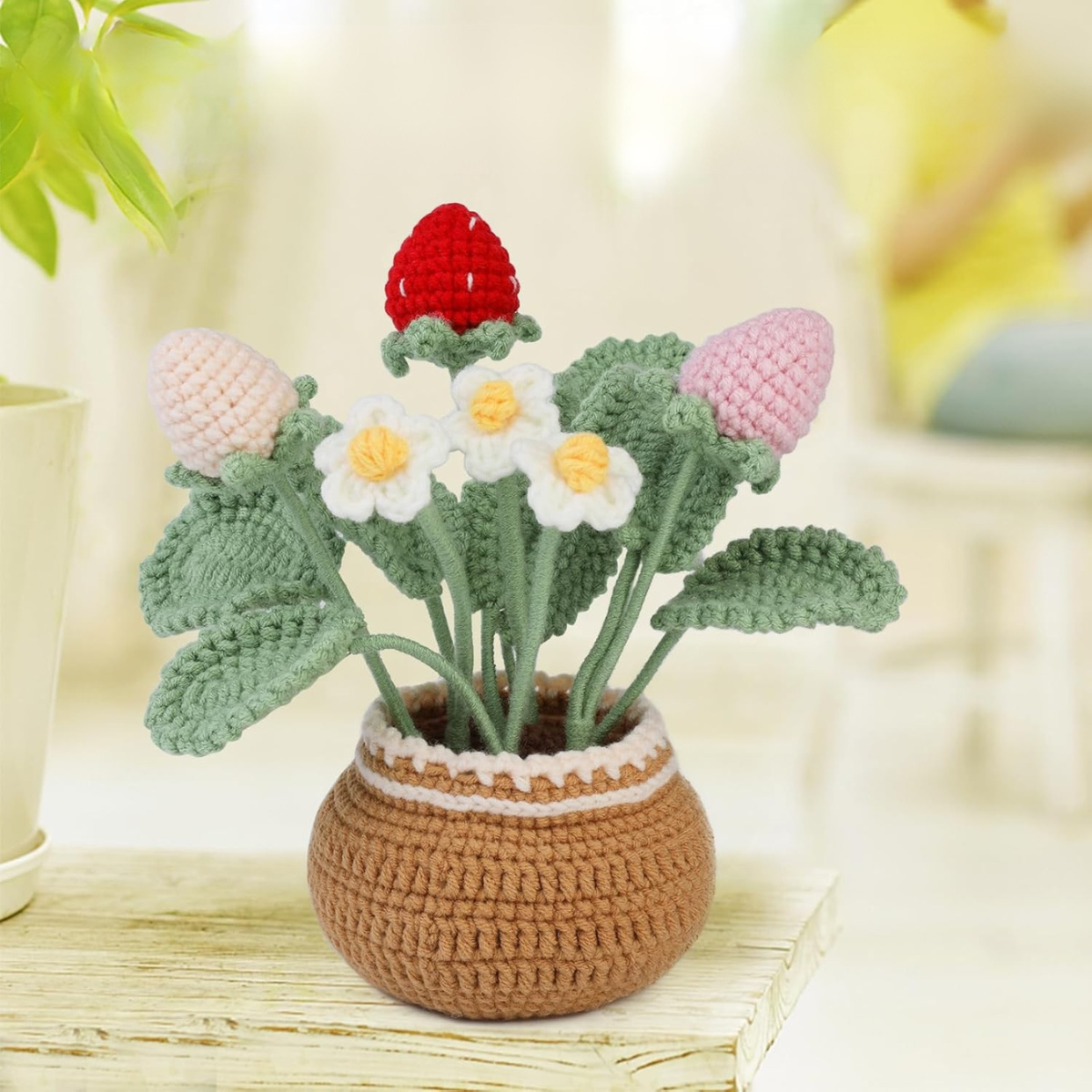 The Crochet Strawberry Plant Kit is perfect for beginners and includes strawberries, white flowers, and green leaves in a brown pot, making a charming addition to any bright indoor table.
