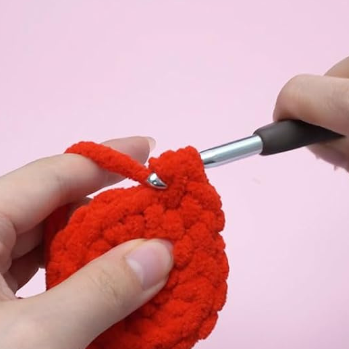 Close-up of hands crocheting with thick red yarn and a metal hook, making the Crochet Heart Plant Kit—a fun DIY home decor project—set against a pale pink background.