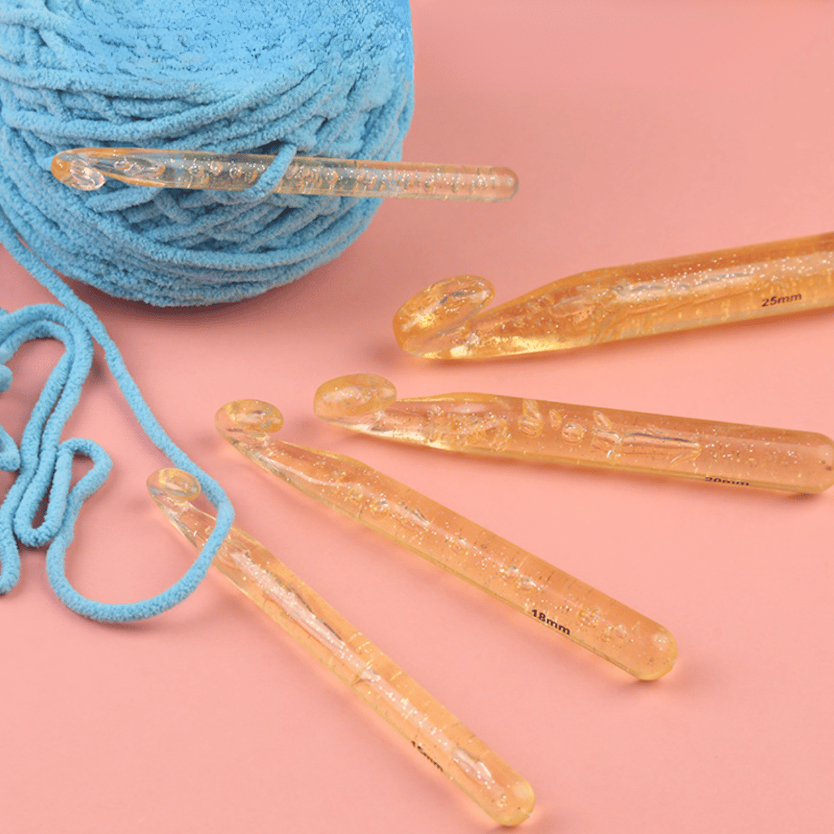 Five translucent amber hooks from the Large Crochet Hooks for Chunky Yarn set and a ball of blue yarn with loose strands are arranged on a pink surface.