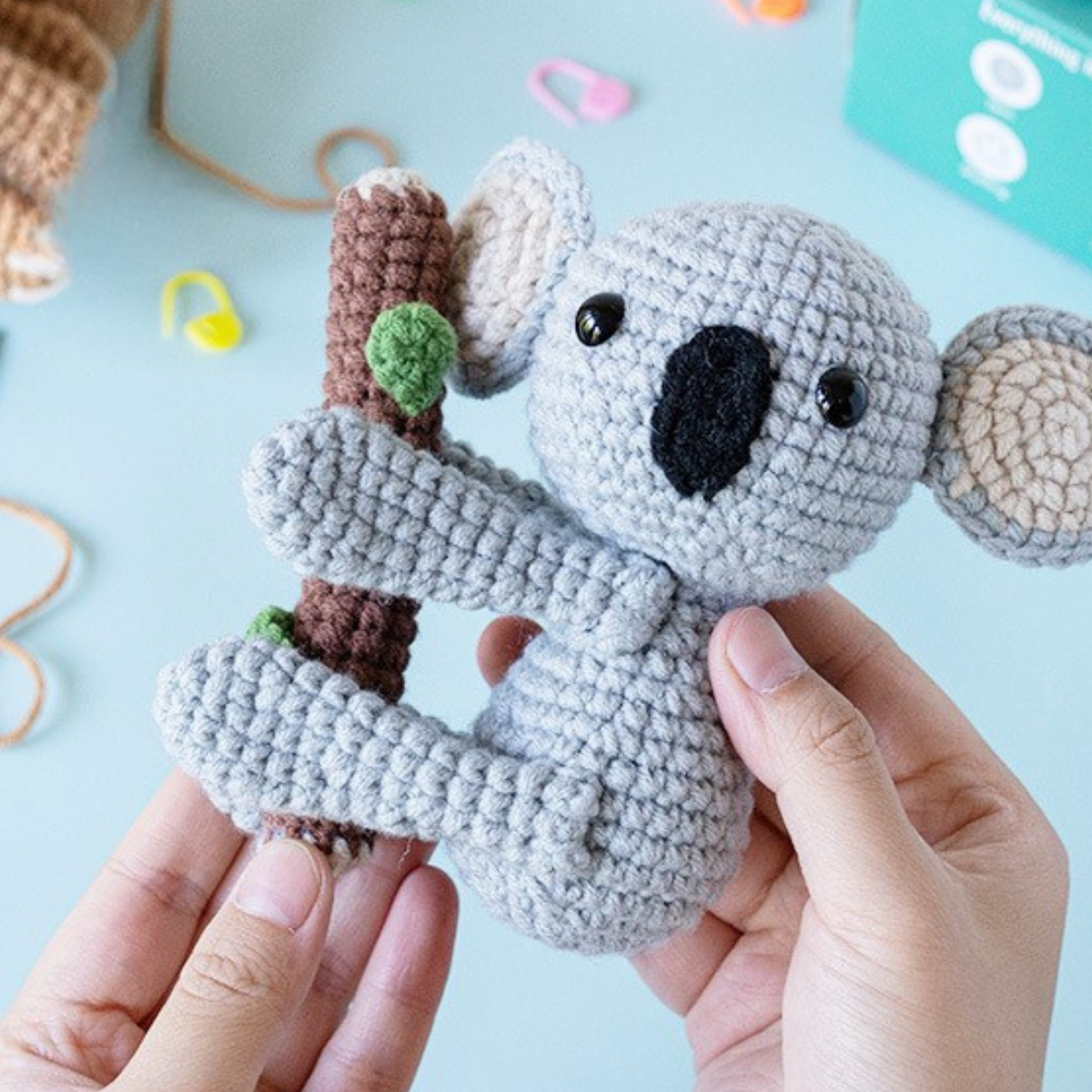 Hands holding a crocheted gray koala hugging a branch, with yarn and crafting supplies in the background—perfect for featuring the Crochet Sloth & Koala Kit.