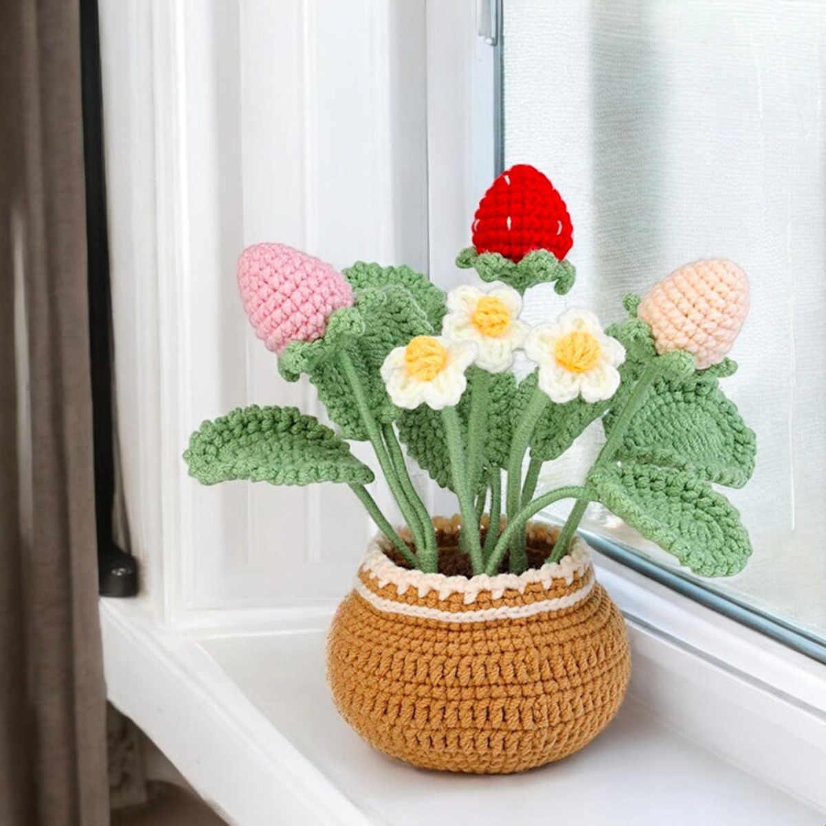 The Crochet Strawberry Plant Kit creates a charming strawberry plant in a brown pot with green leaves, white and yellow flowers, plus pink and red strawberries—perfect decor for any beginner crochet enthusiast.