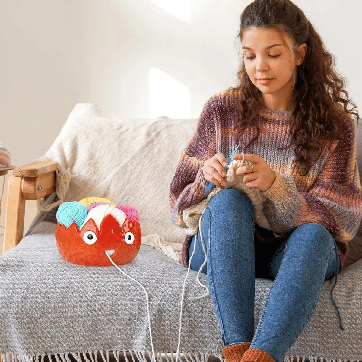 A woman sits on a sofa knitting with beige yarn, which is stored in a colorful plush yarn holder and a Flame Ceramic Yarn Bowl on the couch beside her.