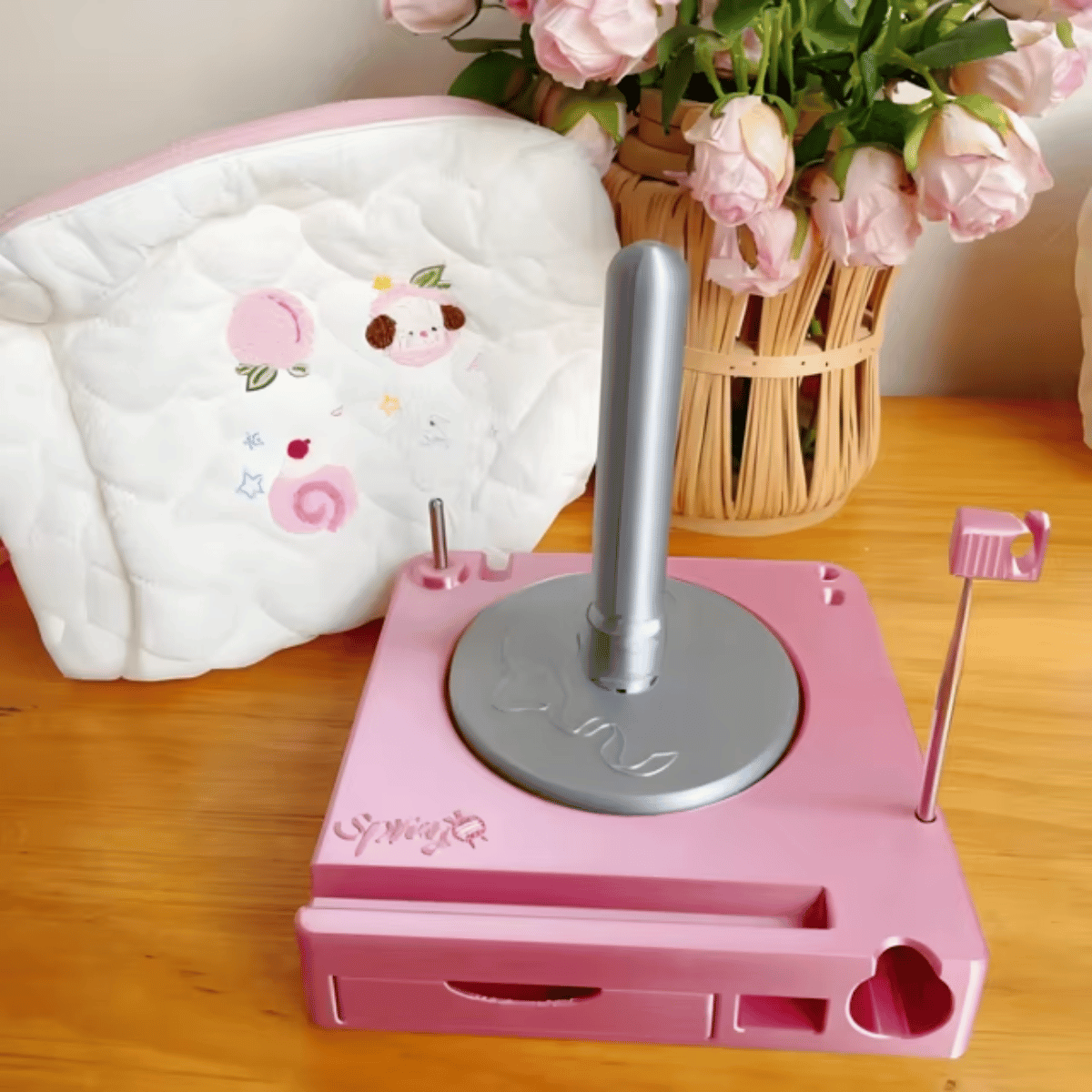A Spinning Yarn Holder with Tool Storage in pink stands on a wooden table beside a white quilted bag and a basket of pink flowers.