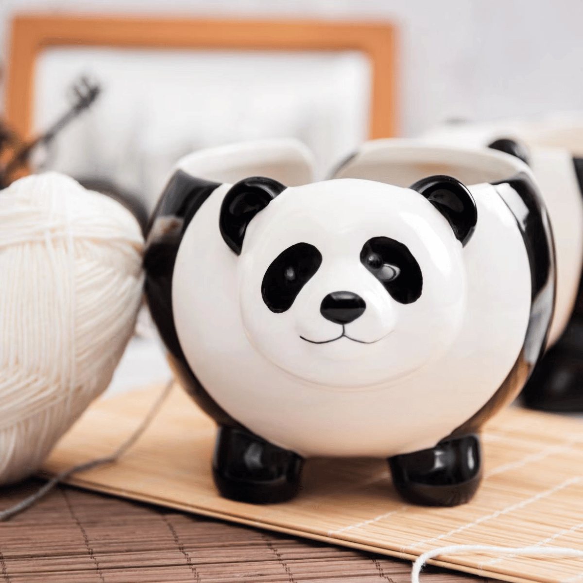 A Panda Ceramic Yarn Bowl sits on a bamboo mat next to an easy-to-use knitting loom and a ball of white yarn, with a framed photo in the background.