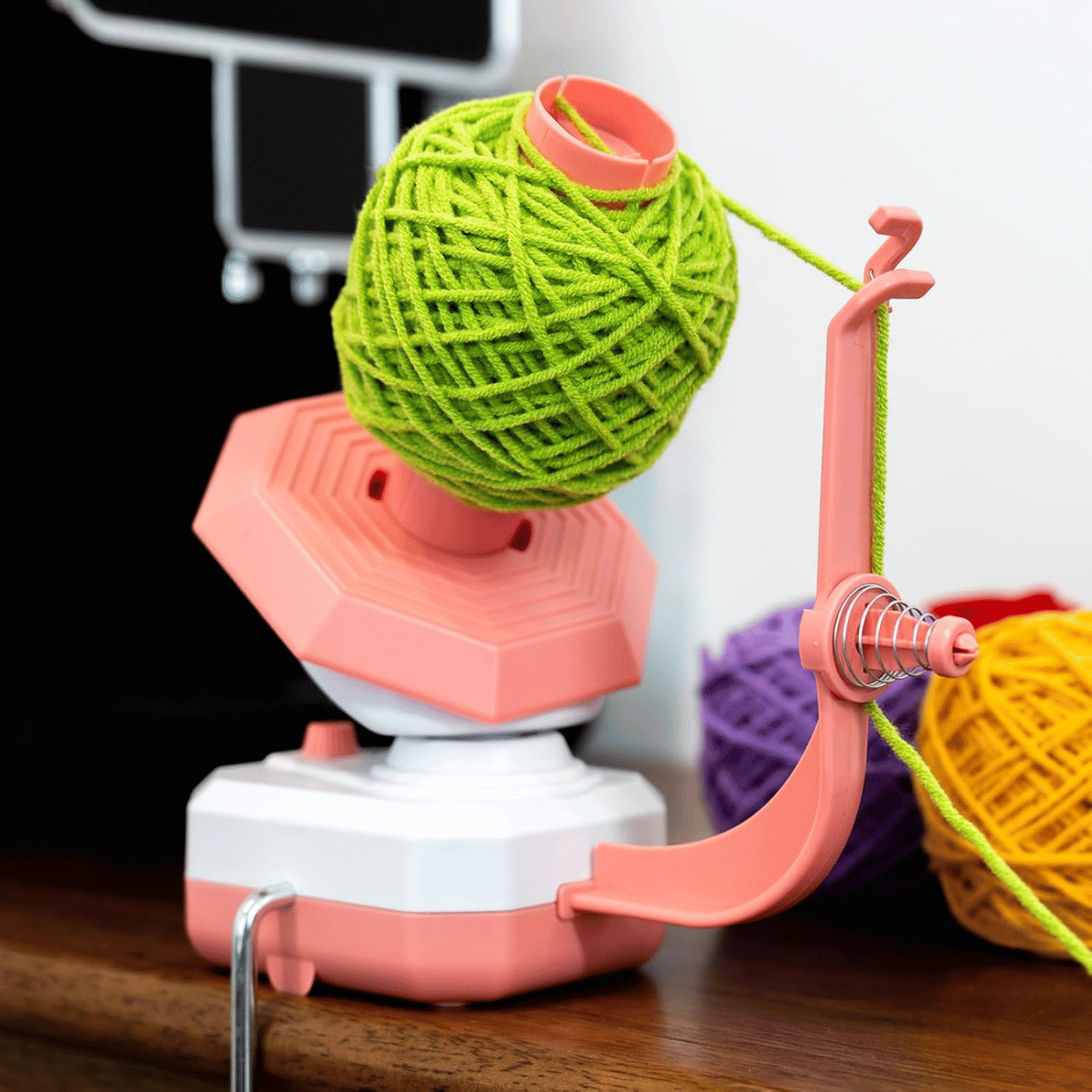 A pink and white Electric Yarn Winder holds a green yarn ball, with yellow and purple yarn balls in the background on a wooden surface.
