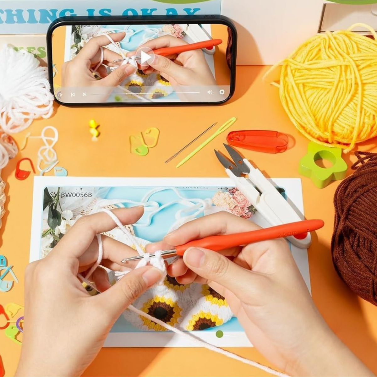 Hands crocheting with white yarn, surrounded by crochet tools, yarn balls, and a phone displaying the Sunflower Crochet Bag Kit tutorial on an orange surface.