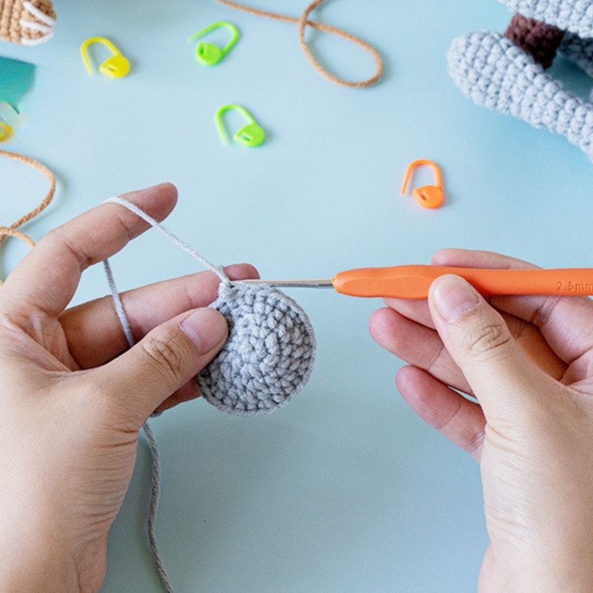 Using the Crochet Sloth & Koala Kit, someone crochets a small, round grey piece with an orange hook while colorful stitch markers rest on the table nearby.
