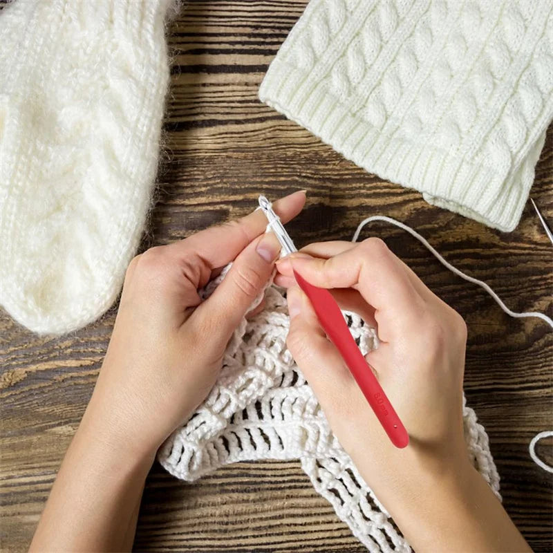 Hands crocheting with a red hook from the Beginner Friendly Crochet Hook Set (31 Pcs with Bag), next to a white knit hat and mitten on a wooden table, perfectly catering to all your crafting needs.