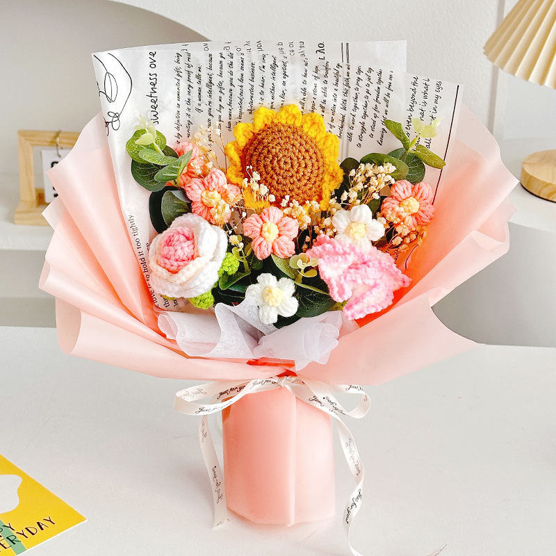 The Crochet Sunflower Bouquet: Blooms That Never Wilt highlights everlasting beauty with a sunflower and pink and white blooms wrapped in pink paper featuring newspaper print. Carefully handcrafted, it perfectly combines artistry and charm.