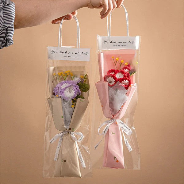 A hand holds up two "Small Bouquet of Crochet Flowers," each wrapped in pink and beige paper, nestled inside clear plastic bags with handles, set against a neutral background.