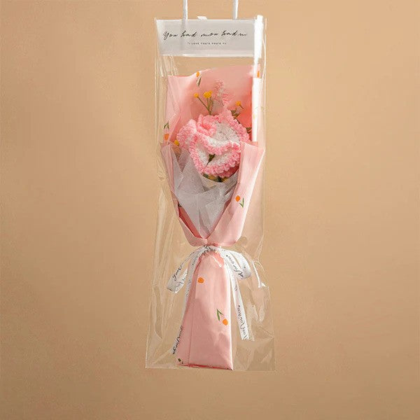A Small Bouquet of Crochet Flowers, expertly handcrafted and wrapped in elegant pink paper and clear plastic, is adorned with a delicate white ribbon against a soft beige background.