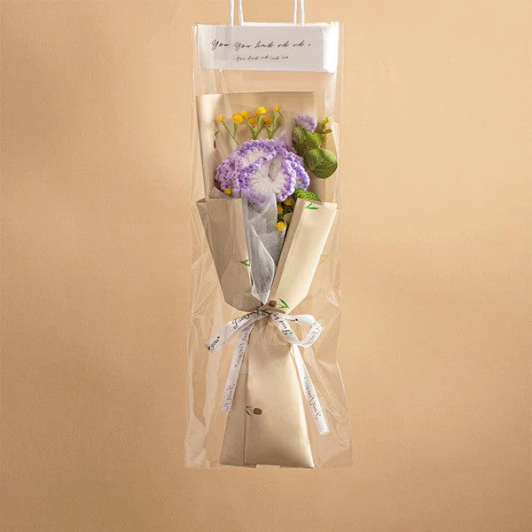 The Small Bouquet of Crochet Flowers is a handcrafted arrangement featuring vibrant purple, yellow, and green crochet blossoms, artistically wrapped in paper and clear plastic, and finished with a delicate white ribbon around the stems.