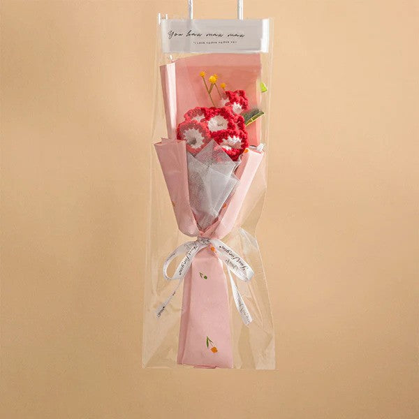 A handcrafted Small Bouquet of Crochet Flowers in pink and red, elegantly wrapped with decorative paper and a ribbon, is displayed against a beige background.
