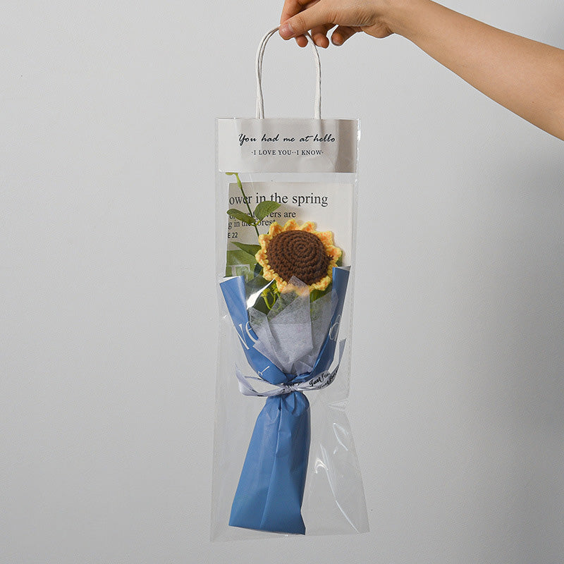 A hand holding a clear gift bag containing a Small Bouquet of Crochet Flowers, featuring an artificial sunflower wrapped in blue paper, with text on the bag.