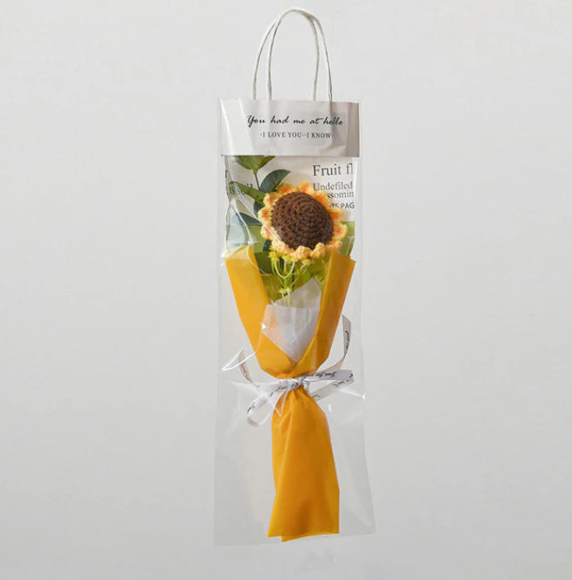 A single handcrafted crochet sunflower, elegantly wrapped in yellow paper, sits inside a clear gift bag with a handle. A thoughtfully attached card at the top adds a personal touch to this Small Bouquet of Crochet Flowers.