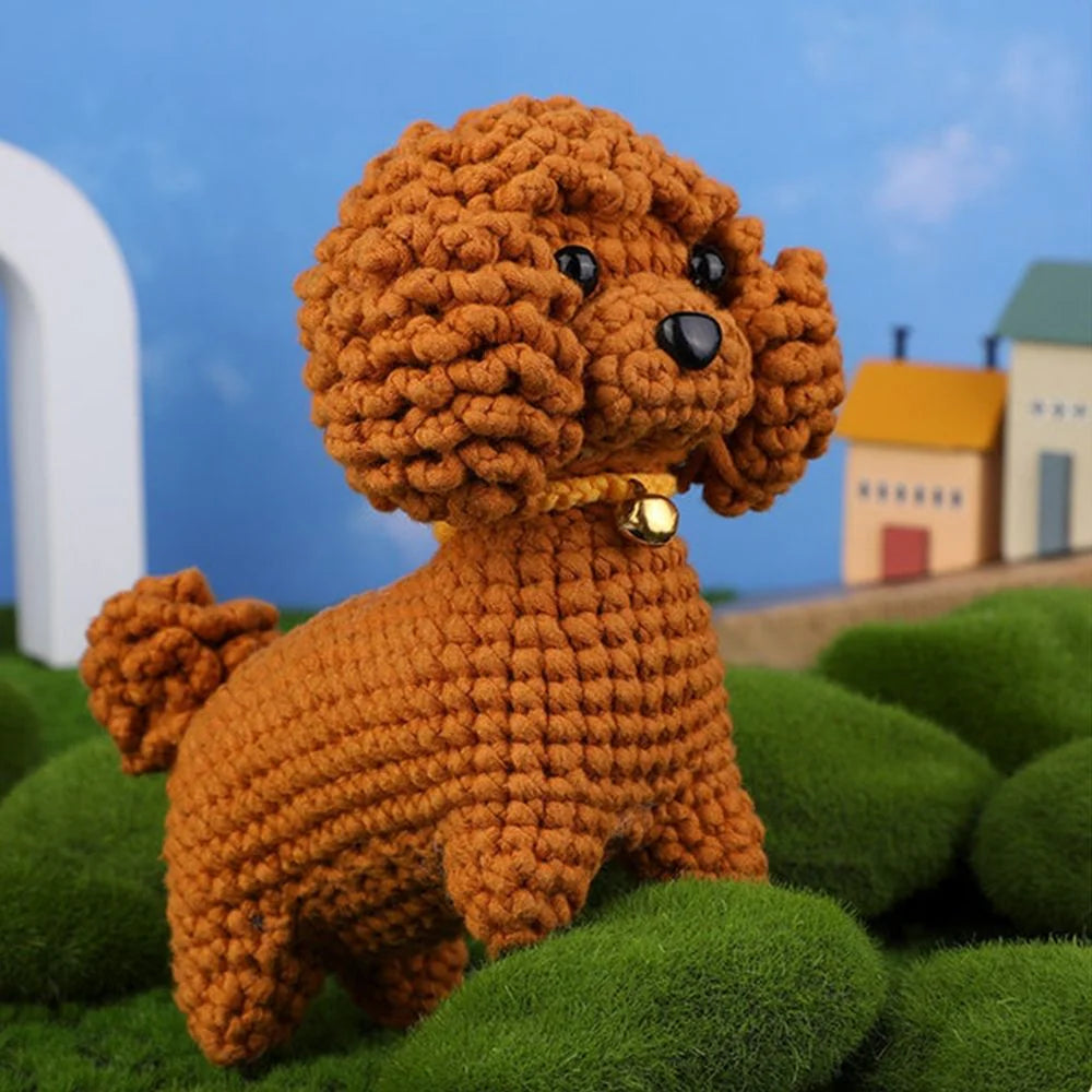 The Crochet Poodle Kit makes a handmade poodle plush with curly brown texture, a yellow collar with bell, standing on green plush, against a blue sky and small buildings background.