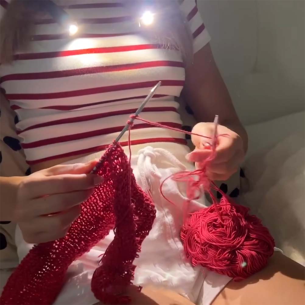 A person, dressed in a striped shirt, is using a Crocheting/Knitting Neck Lamp to knit a red fabric with red yarn, while skillfully maneuvering two knitting needles.