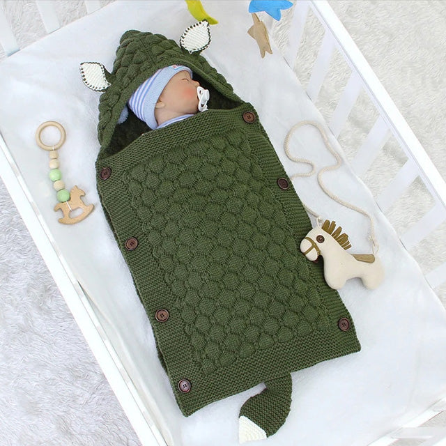 A baby sleeps peacefully in a crib, nestled in the cozy comfort of a Knitted Baby Sleeping Bag Wrap Blanket (bundle) in green, adorned with cute animal ears and a tail. Beside them are a few toys, including an adorably designed wooden horse and rattle.