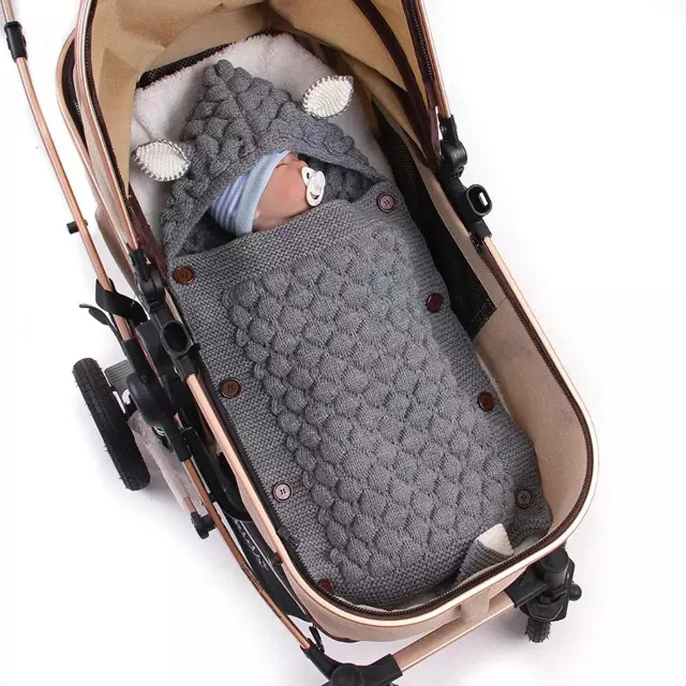 A baby in a stroller, snug in a Knitted Baby Sleeping Bag Wrap Blanket bundle that boasts an adorable design, wrapped in a gray, textured knit blanket with ear-like features on the hood.