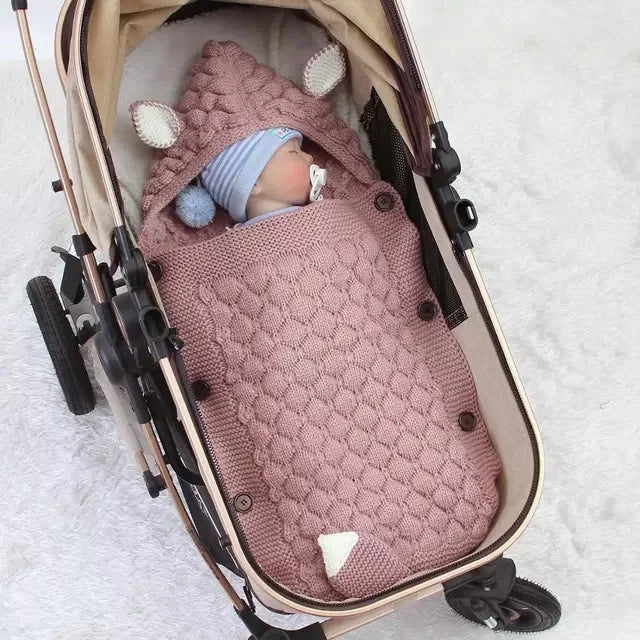 Wrapped in the Knitted Baby Sleeping Bag Wrap Blanket, featuring a charming pink design with bear ears, a baby sits adorably in a stroller and wears a blue hat.
