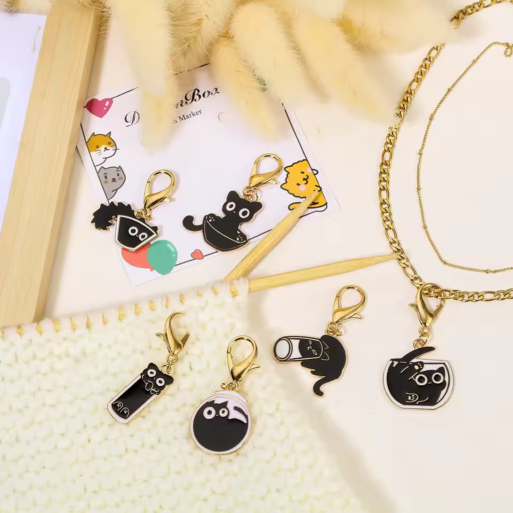 The Elegant Black Cat Stitch Markers Set (6pcs) is displayed on cream knit fabric with decorative postcards and beige dried flowers, making it a charming knitting accessory or thoughtful gift.