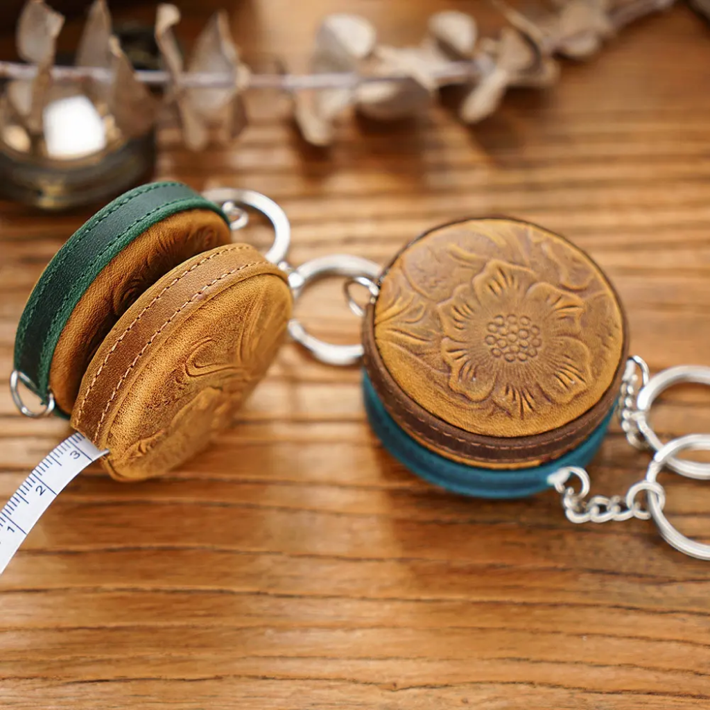 Two Mini Leather Tape Measure 60" cases, round and handmade with floral embossing and keychains, are displayed on a wooden surface—perfect as vintage sewing accessories.