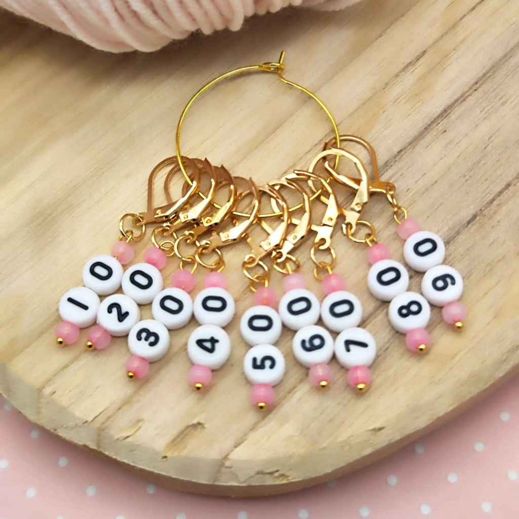 The Acrylic Numbered (10-90) Stitch Marker Set 9Pcs features acrylic markers with gold details and pink/white beads, numbered in tens from 10 to 90, perfect for knitting or crochet yarn crafts and displayed on a wooden surface.