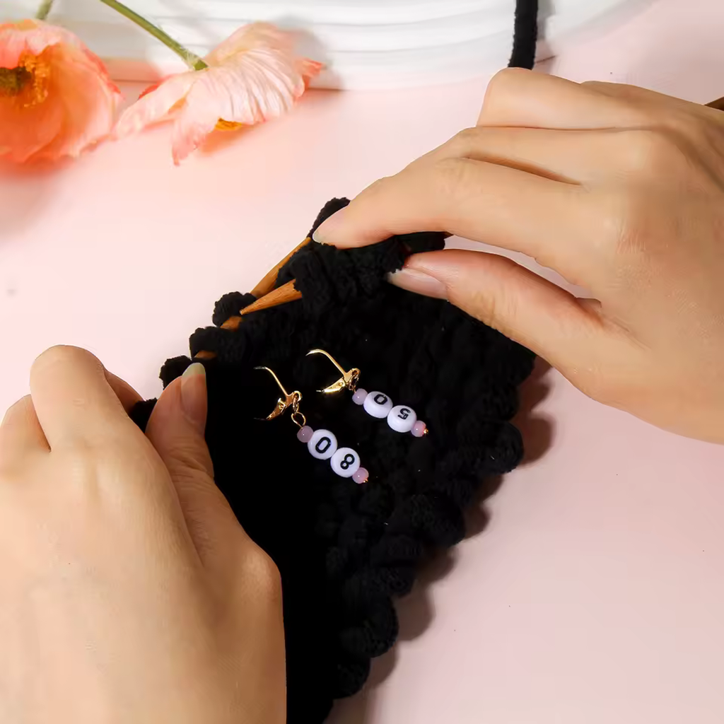 Two hands knitting with black yarn using wooden needles, featuring the Acrylic Numbered (10-90) Stitch Marker Set 9Pcs—ideal for knitting and crochet lovers. Beaded stitch marker earrings and orange flowers complete this charming yarn craft scene.