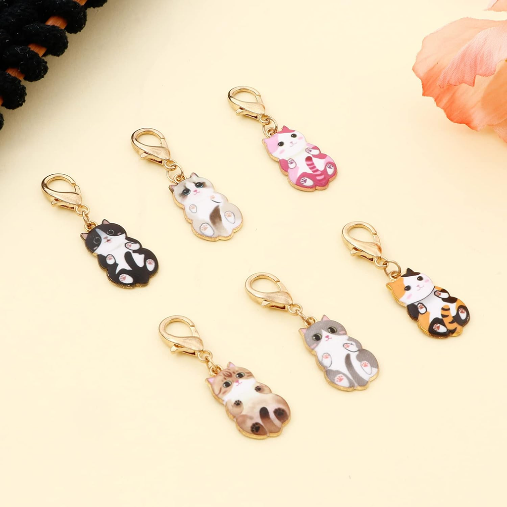 The Enamel Cat Shape Stitch Marker Set 6Pcs features gold lobster clasps and comes arranged on a light yellow surface near a peach-colored flower—ideal for crochet projects and craft lovers.