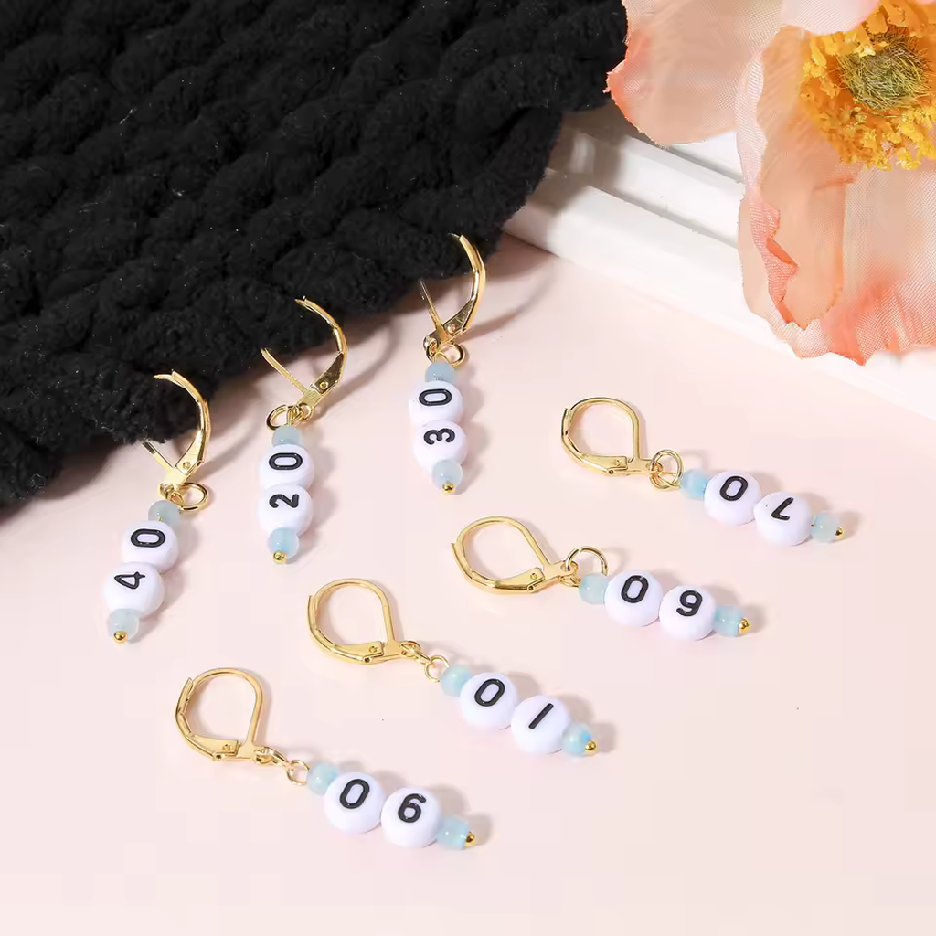 The Acrylic Numbered (10-90) Stitch Marker Set 9Pcs features gold leverback markers for knitting and crochet, each with a beaded number, perfect for yarn crafts. Shown on pink with a knitted fabric and peach flower.