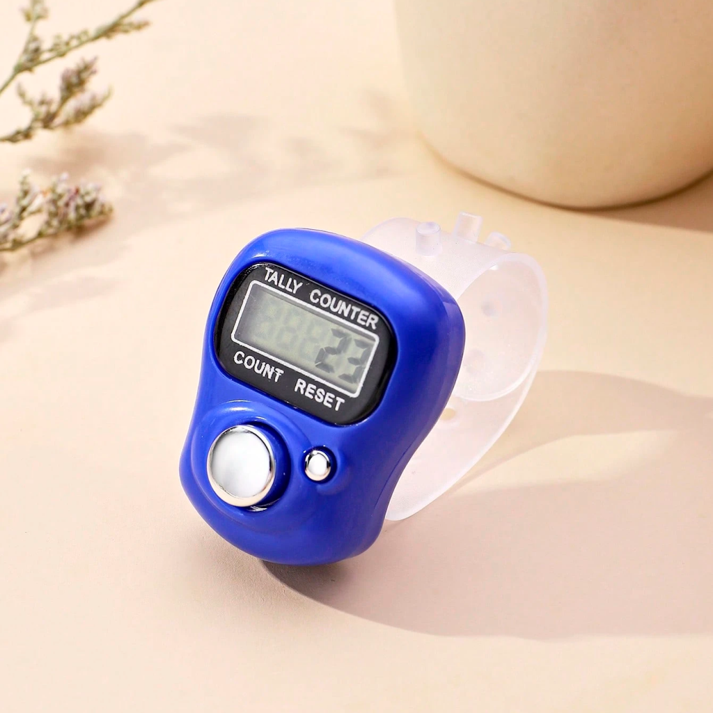 The Digital Row Counter, featuring a blue display screen and button on a clear adjustable strap, rests on a light surface beside a plant—perfect for easy tracking during crafts.