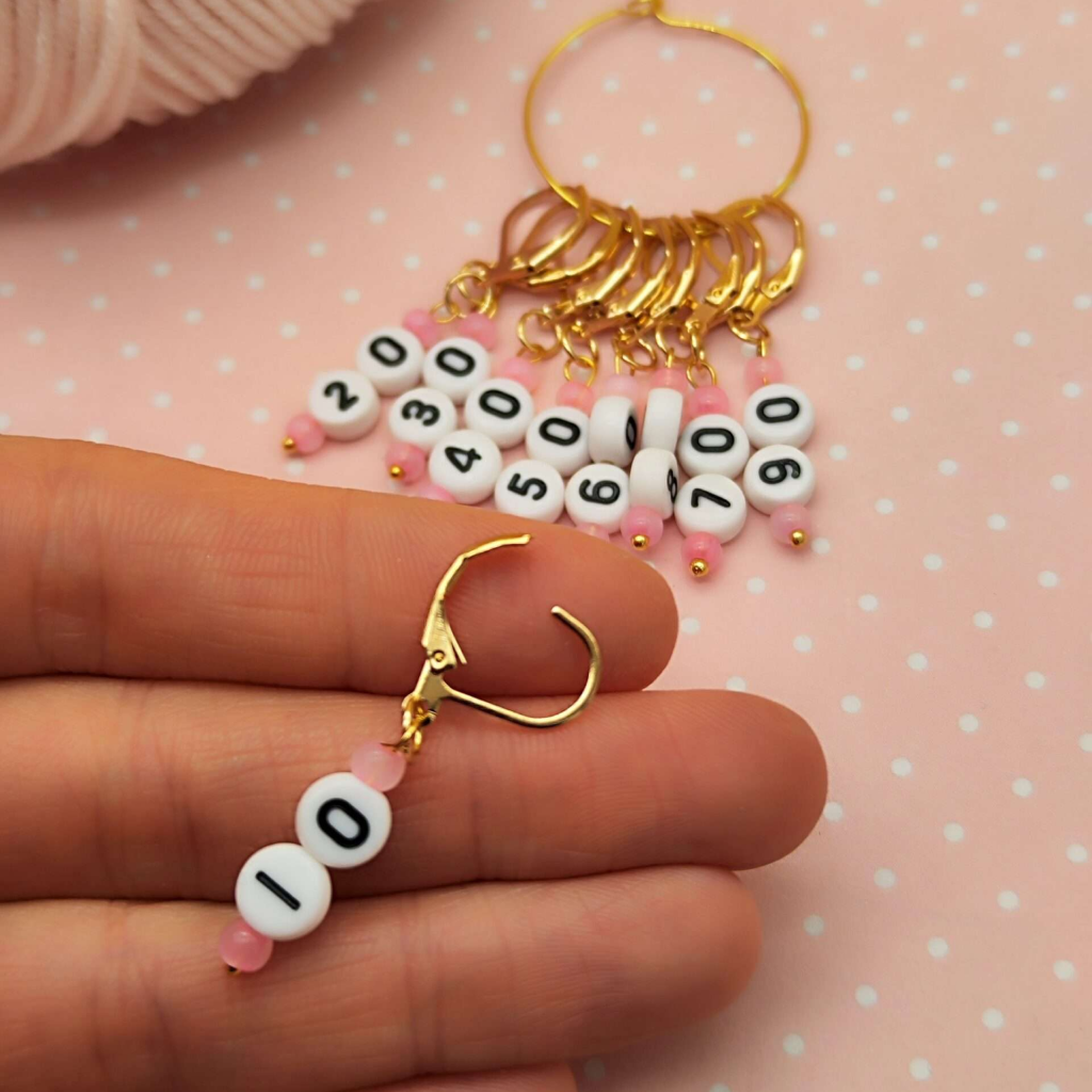 A hand holds an Acrylic Numbered (10-90) Stitch Marker from the 9-piece set, ideal for knitting and crochet. More markers with numbers 10–90 are displayed on a gold ring in the background—perfect accessories for yarn crafts.