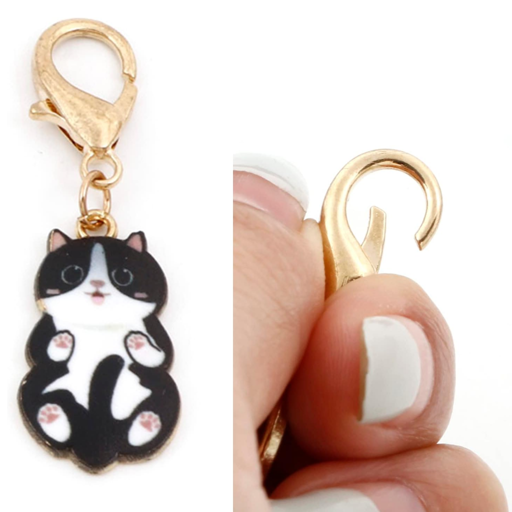 Close-up of a gold keychain clasp holding a black and white charm from the Enamel Cat Shape Stitch Marker Set 6Pcs, ideal for crafting or as enamel cat stitch markers; a hand with white nail polish is opening the clasp.