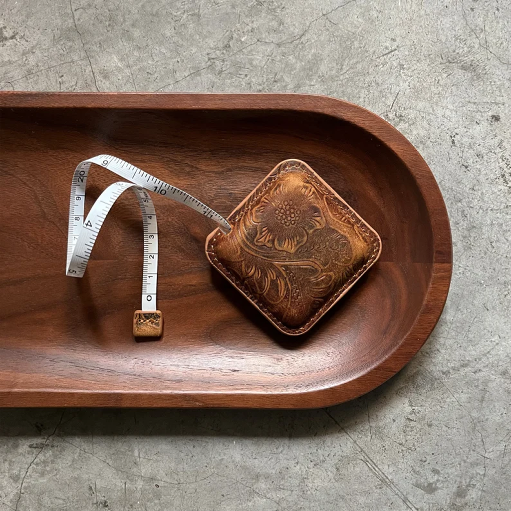 The Mini Leather Tape Measure 60", featuring vintage floral embossing in brown, is displayed on a wooden tray with its white tape extended—a chic tool for knitting and crocheting projects on a concrete surface.