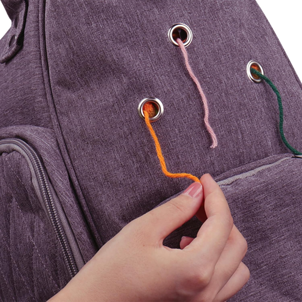 A hand pulls an orange yarn strand through a metal grommet on the Knitting and Crochet Organizer Backpack, made of durable 600D Oxford fabric in purple, with pink and green yarn strands threaded through nearby grommets.