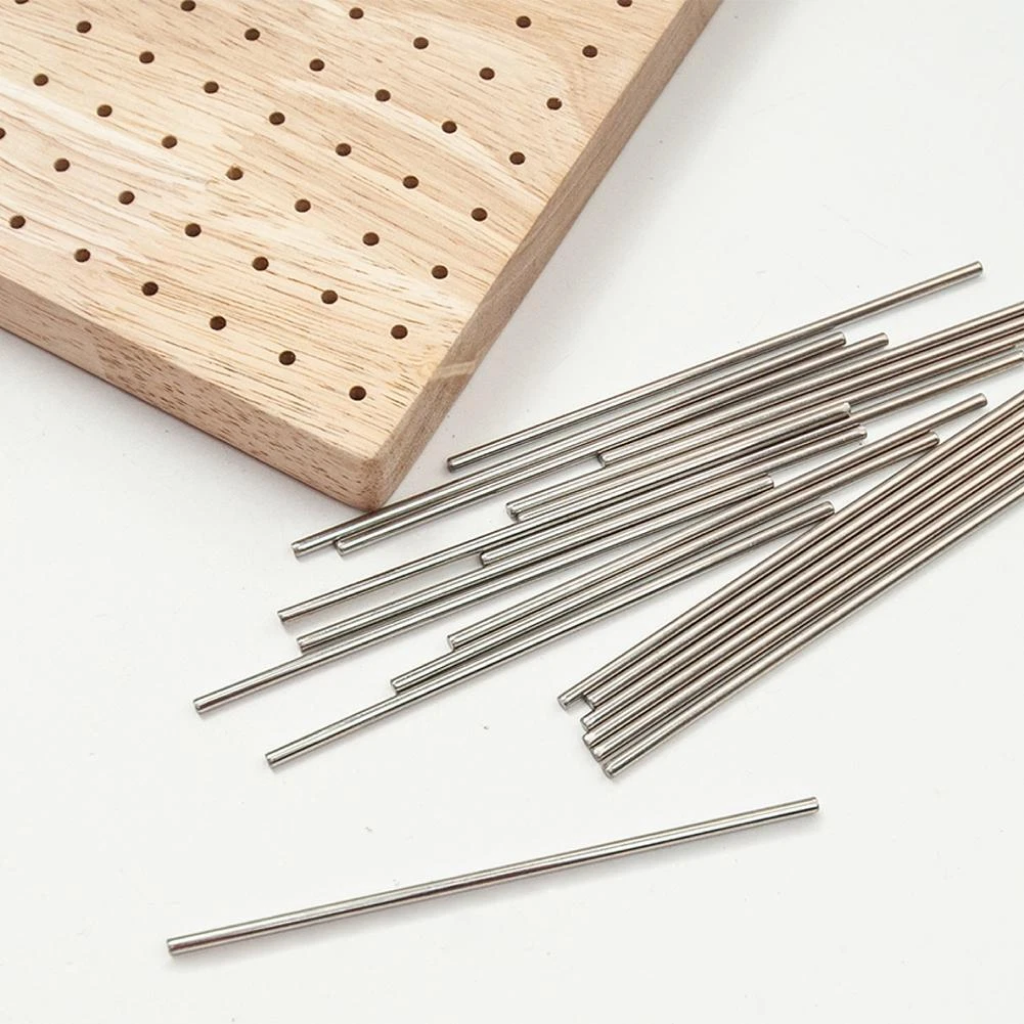 The Crochet Blocking Board, a wooden board with evenly spaced holes, is displayed beside several metal rods on a white surface—ideal for organizing your crochet projects.