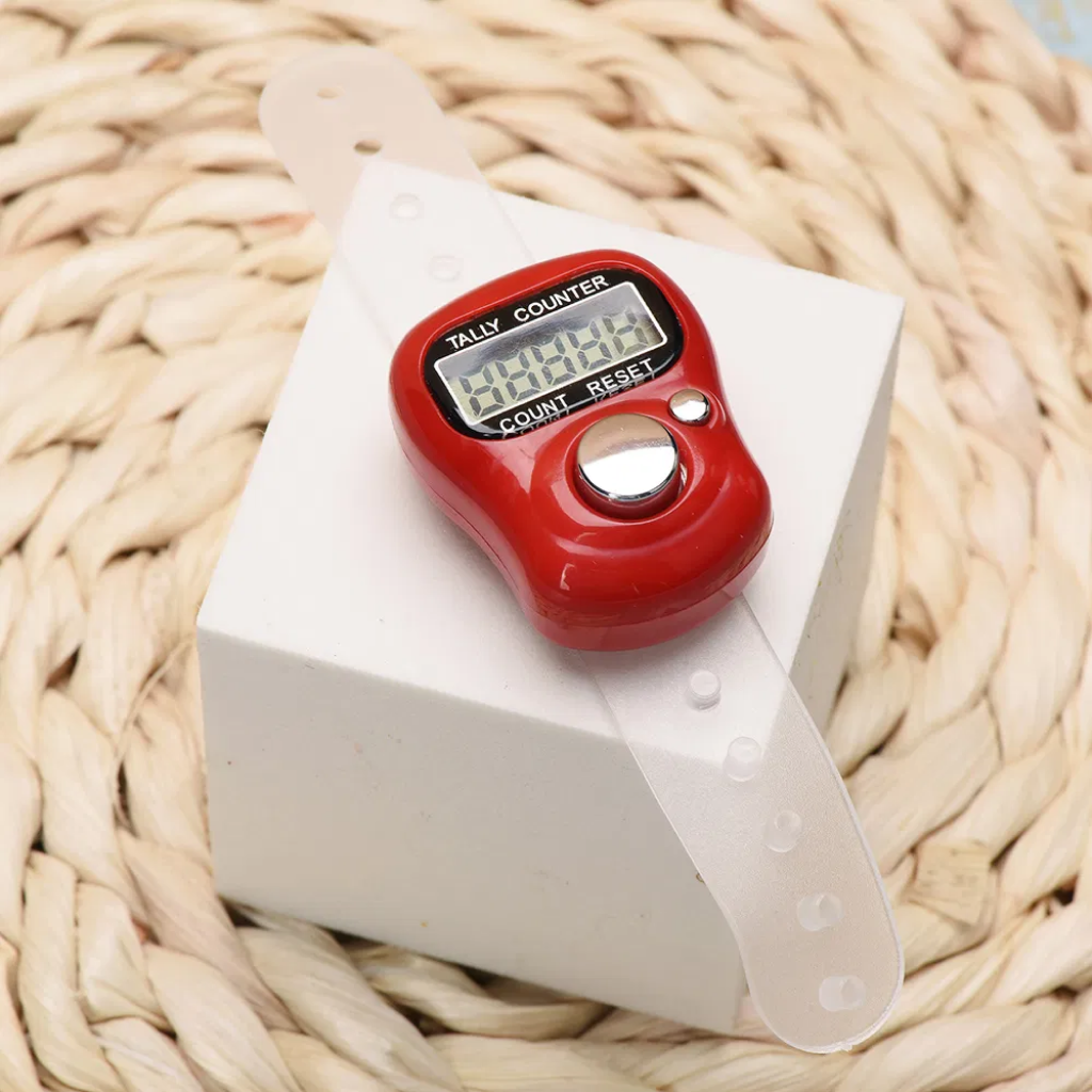 The Digital Row Counter, featuring a red body and transparent adjustable strap, sits on a white box atop a woven surface—offering simple tracking with its compact, portable design.