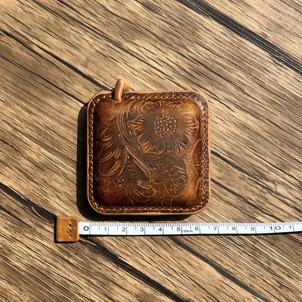 A square Mini Leather Tape Measure 60" with floral embossing and vintage charm sits on a wood surface next to a tape showing its approximately 9cm width—ideal for knitting and crocheting projects.