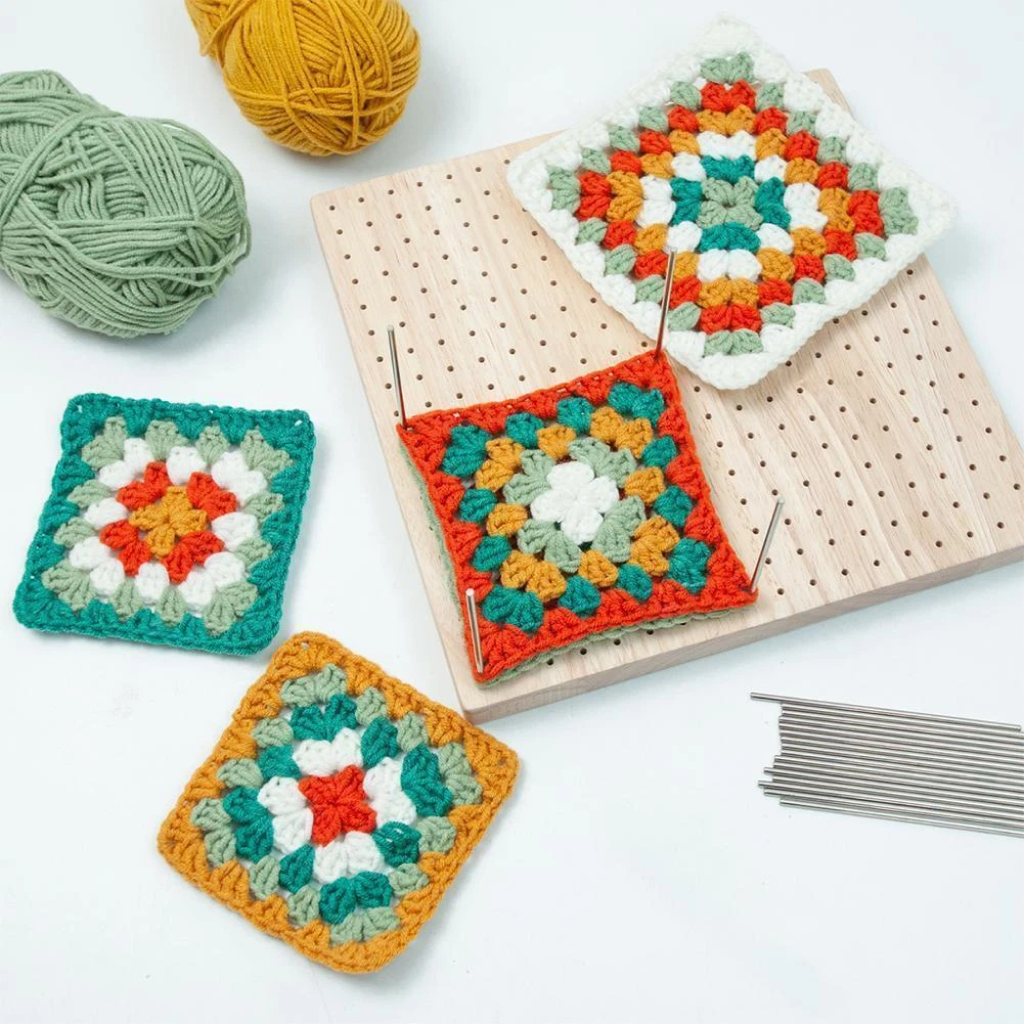 Four colorful crochet granny squares and yarn balls are arranged beside the Crochet Blocking Board, which features pins and metal rods—ideal for finishing and perfecting your crochet projects.