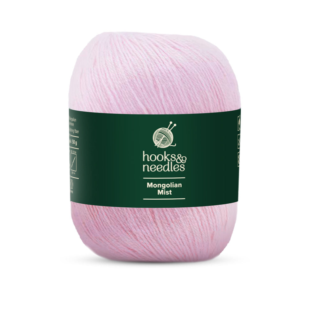 A skein of light pink "Mongolian Mist Yarn 50g" presented on a dark green band with the label "Hooks & Needles," made from super fine-weight yarn for a smooth, luxurious feel.
