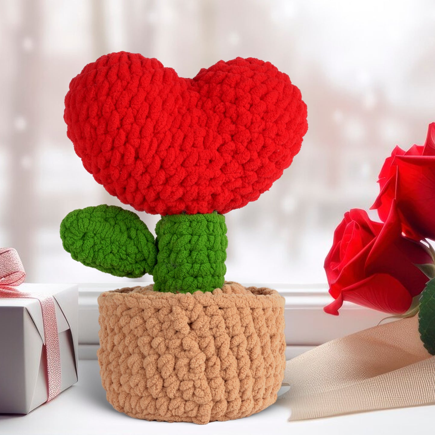 The Crochet Heart Plant Kit features a red heart-shaped top, green stem and leaves in a beige pot. Gift box and red roses in the background make it ideal DIY crochet decor or a charming gift.