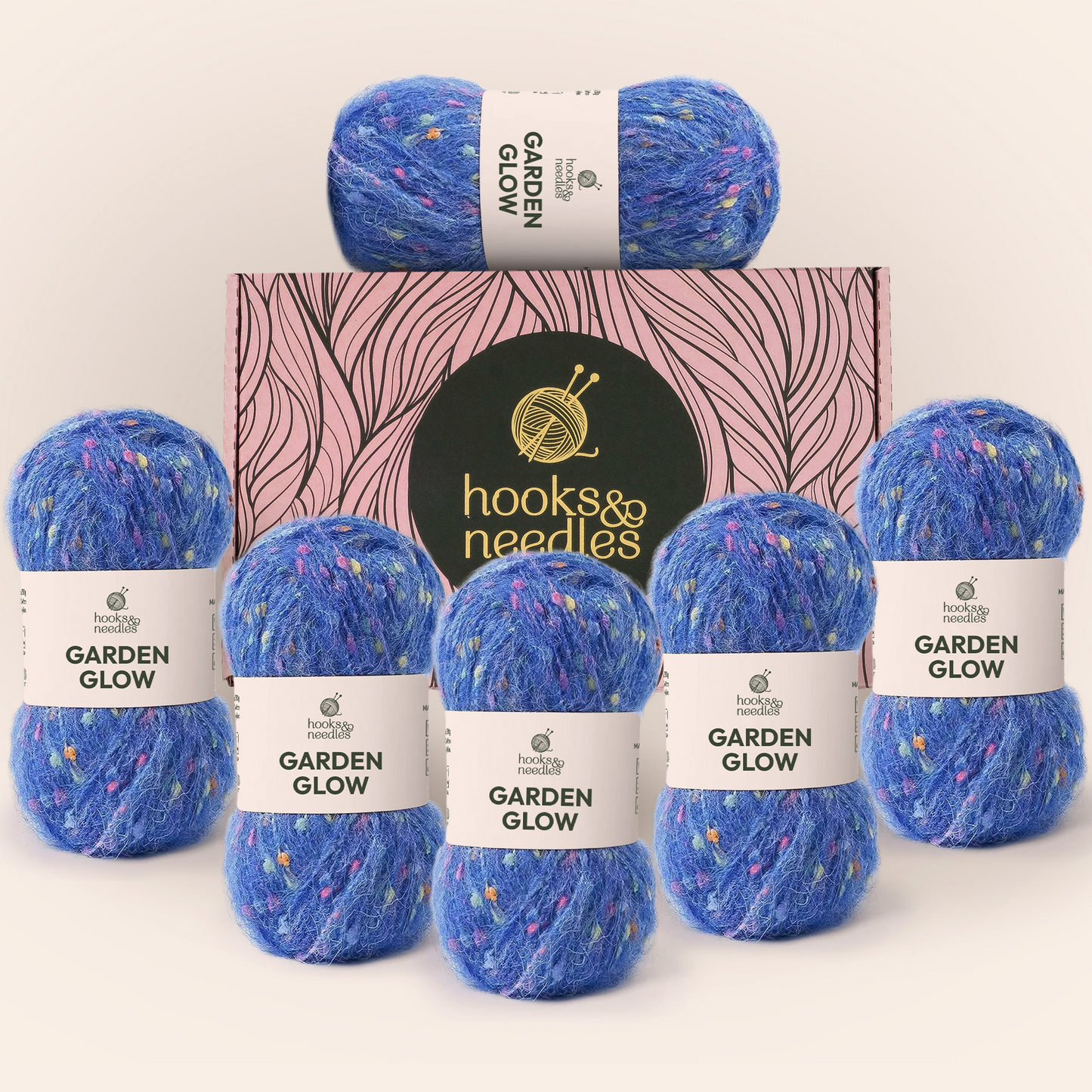 Six skeins of blue speckled Garden Glow yarn are arranged in front of a pink Hooks & Needles Yarn Subscription Box featuring a yarn ball and knitting needles logo.