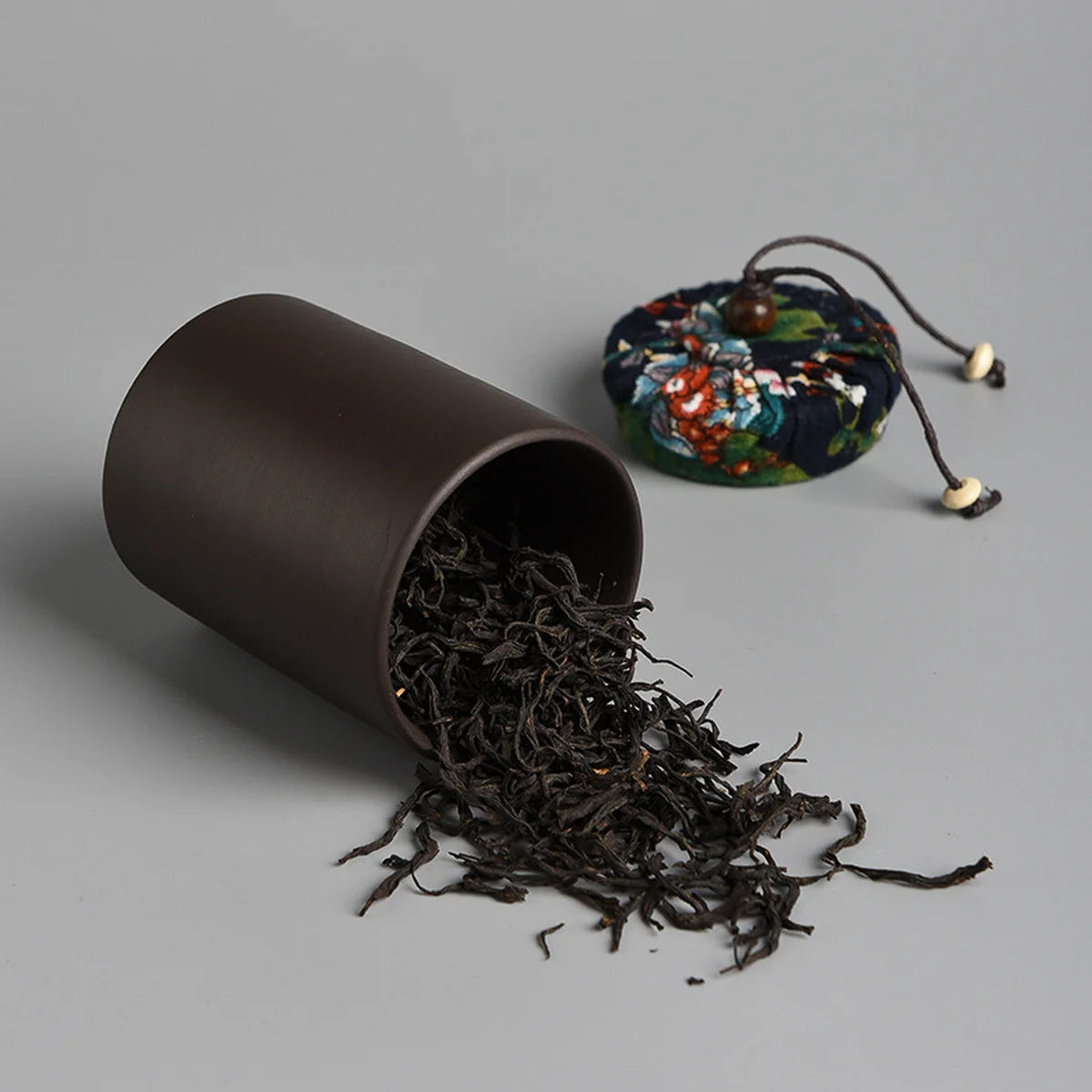 A dark ceramic cup tipped over with loose black tea leaves scattered, beside a round, floral-patterned fabric pouch with a drawstring—ideal for the Tea Set with Case (Bundle24), perfect for on-the-go brewing.