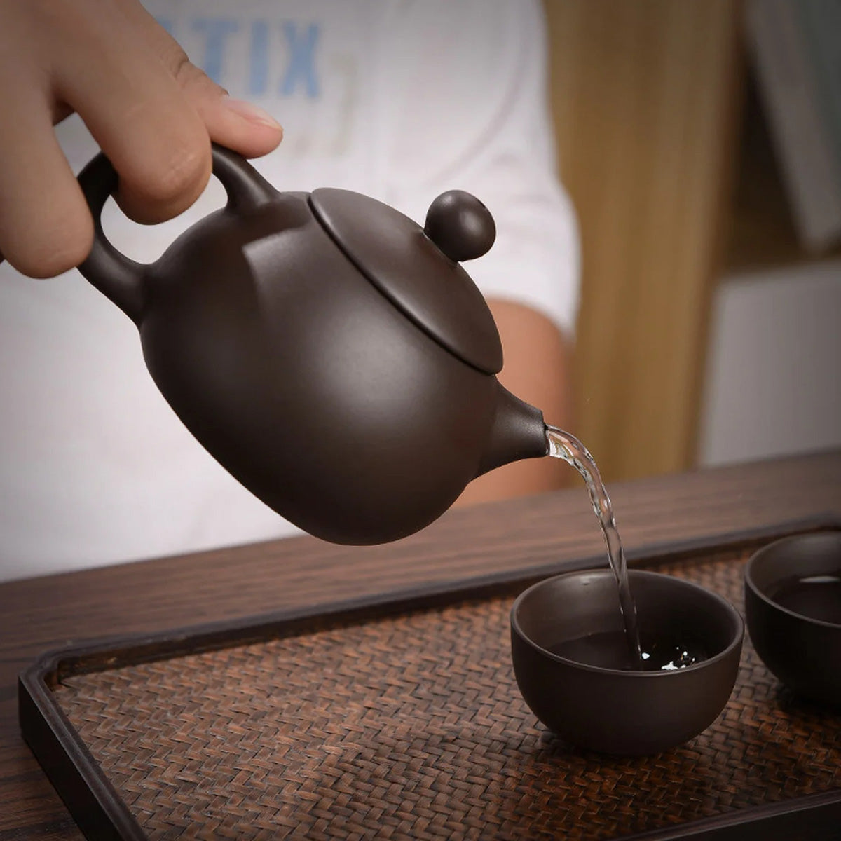 A person pours tea from a dark teapot into small matching cups on a woven mat, all part of the Tea Set with Case (Bundle24), ideal for travel.