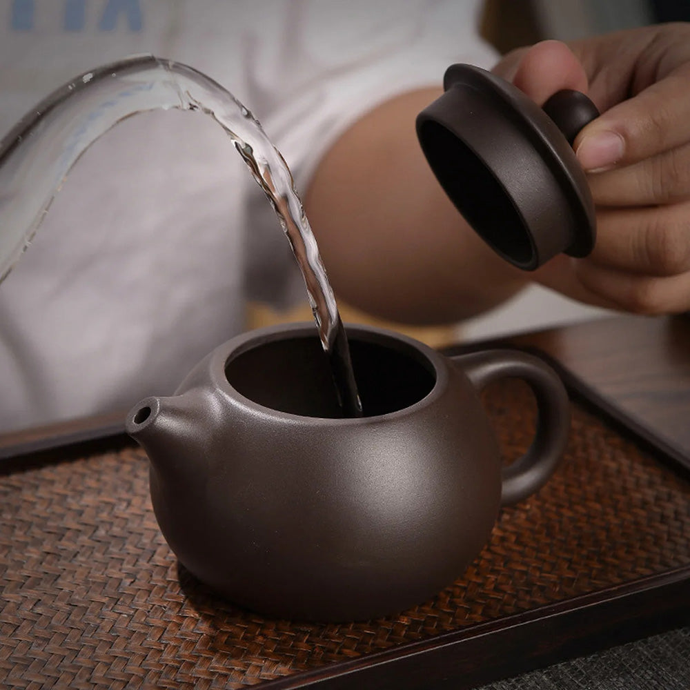 A person pours water into a dark brown teapot from the Tea Set with Case (Bundle24), with its lid removed, placed on a woven mat.