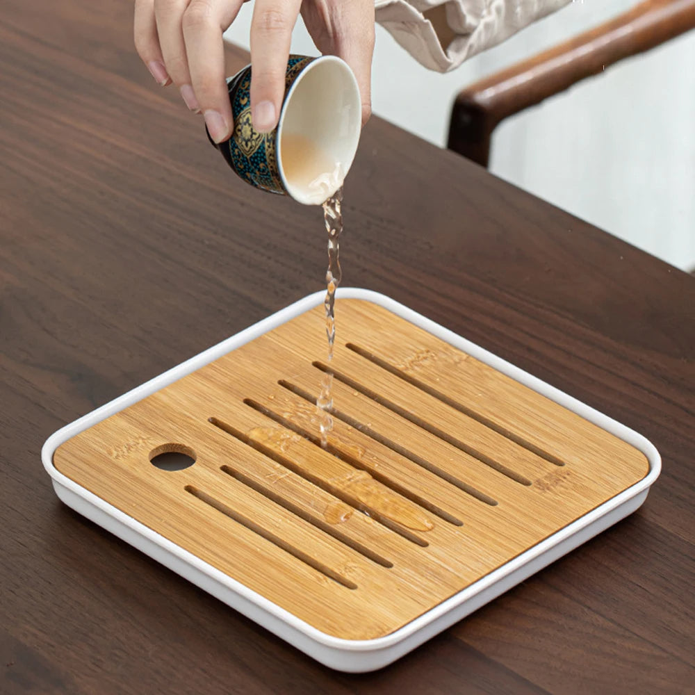 A tea connoisseur carefully pours liquid from the Tea Making Set With Cups and Case onto a square wooden slatted tray resting on a dark wooden table.
