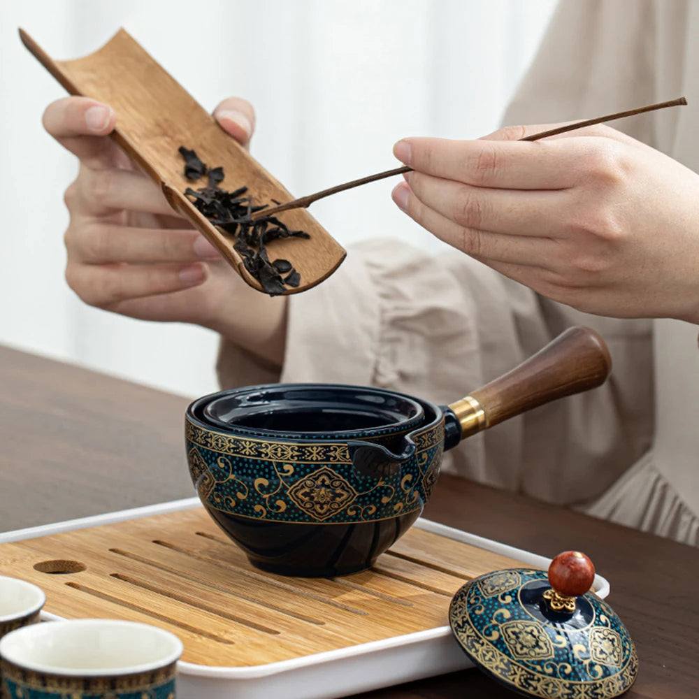 A person carefully uses a wooden scoop to add tea leaves into a decorative teapot, which is part of the exquisite Tea Making Set With Cups and Case on a tray, with cups thoughtfully arranged nearby, making it perfect for seasoned tea connoisseurs.