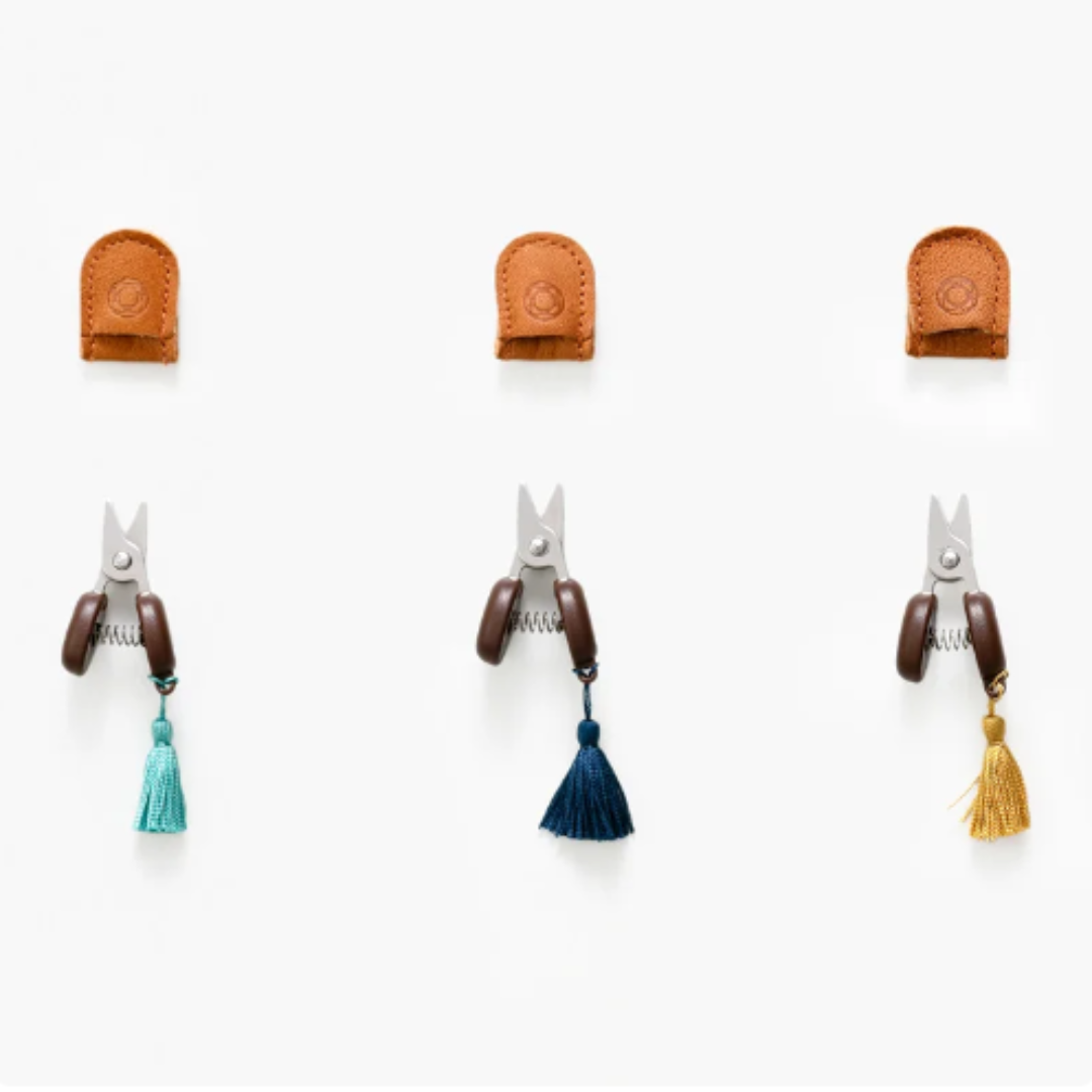 Hidden beneath brown leather sheaths are three small scissors, featuring dark handles adorned with vibrant turquoise, blue, and gold tassels. These compact tools offer perfect portability and precision craftsmanship.