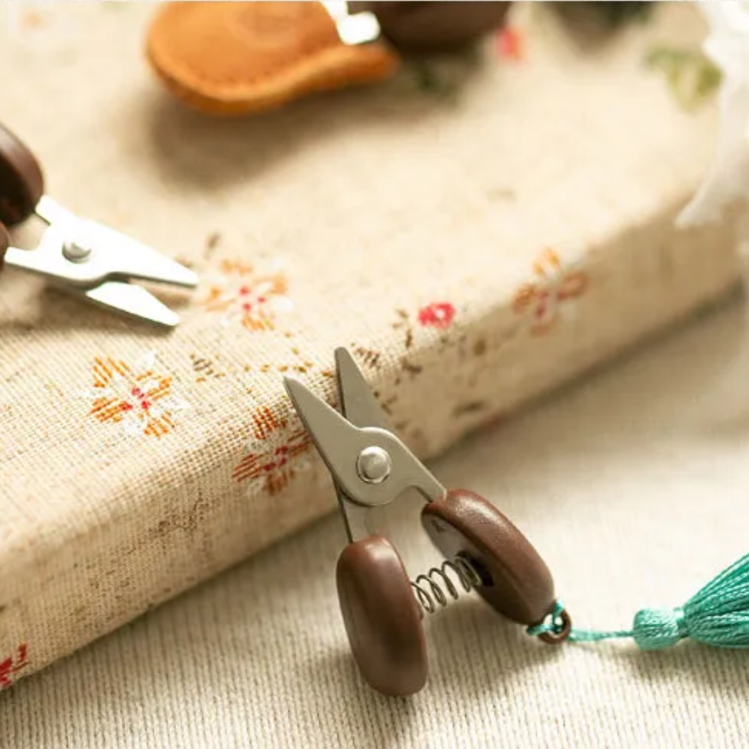 A pair of Small Scissors, known for their compact and portable design with wooden handles, rests on a floral-patterned book, providing precision for delicate crafts.